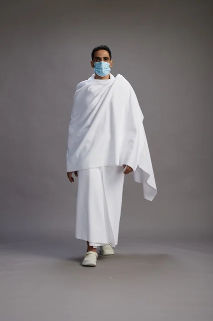 An Arab Saudi Gulf man in Ihram clothing wearing a mask standing ready for Hajj, protection from the coronavirus, full body, gray background.