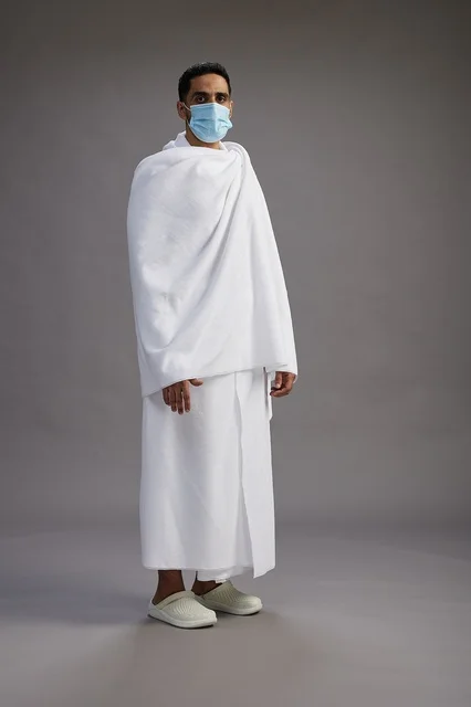 رجل عربي  سعودي  خليجي بملابس الإحرام يرتدي كمامة واقف استعدادًا للحج, الوقايه من فايروس كورونا, كامل الجسم, خلفيه رماديه