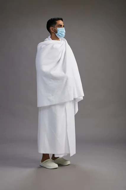 رجل عربي  سعودي  خليجي بملابس الإحرام يرتدي كمامة واقف استعدادًا للحج, الوقايه من فايروس كورونا, كامل الجسم, خلفيه رماديه