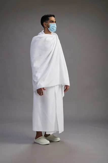 An Arab Saudi Gulf man in Ihram clothing wearing a mask standing ready for Hajj, protection from the coronavirus, full body, gray background.