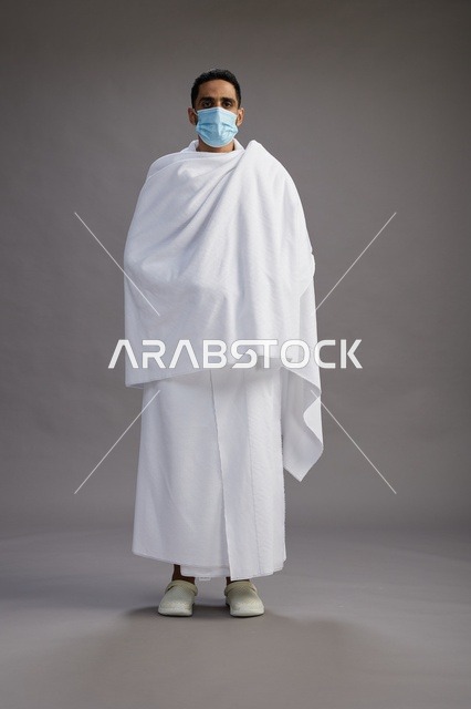 رجل عربي  سعودي  خليجي بملابس الإحرام يرتدي كمامة واقف استعدادًا للحج, الوقايه من فايروس كورونا, كامل الجسم, خلفيه رماديه