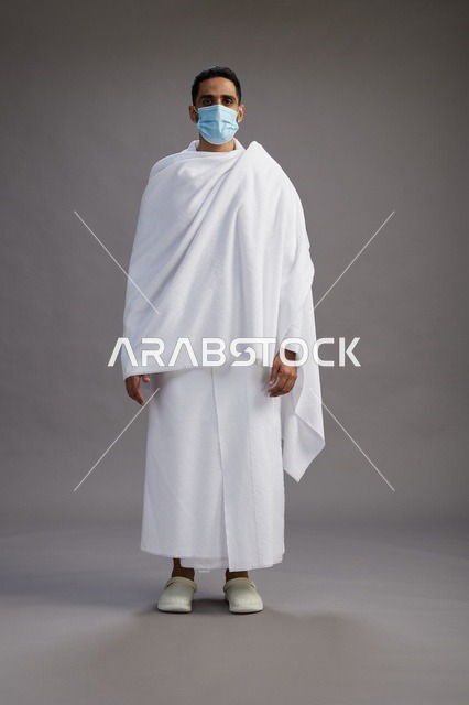 رجل عربي  سعودي  خليجي بملابس الإحرام يرتدي كمامة واقف استعدادًا للحج, الوقايه من فايروس كورونا, كامل الجسم, خلفيه رماديه