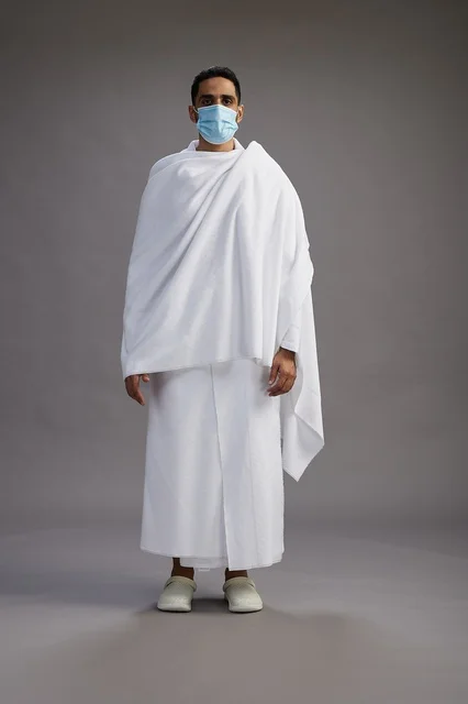 رجل عربي  سعودي  خليجي بملابس الإحرام يرتدي كمامة واقف استعدادًا للحج, الوقايه من فايروس كورونا, كامل الجسم, خلفيه رماديه