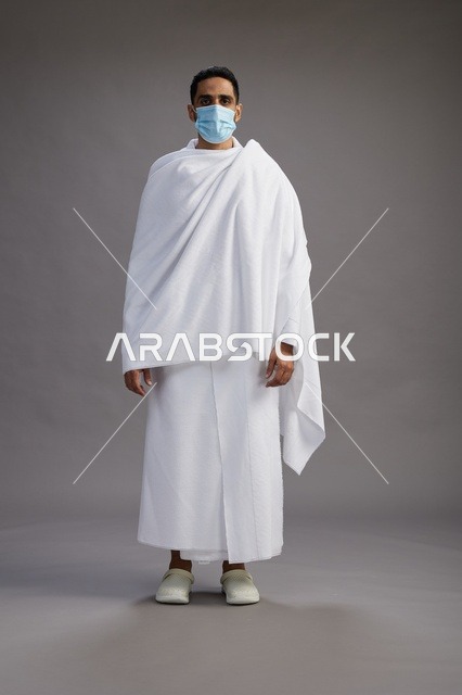 رجل عربي  سعودي  خليجي بملابس الإحرام يرتدي كمامة واقف استعدادًا للحج, الوقايه من فايروس كورونا, كامل الجسم, خلفيه رماديه