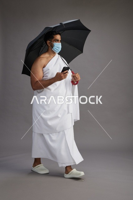 حاج  عربي سعودي خليجي بملابس الإحرام يحمل مظلة ويرتدي كمامة, استخدام الهاتف المحمول , اداء مناسك الحج والعمره, كامل الجسم, خلفيه رماديه