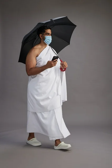 حاج  عربي سعودي خليجي بملابس الإحرام يحمل مظلة ويرتدي كمامة, استخدام الهاتف المحمول , اداء مناسك الحج والعمره, كامل الجسم, خلفيه رماديه