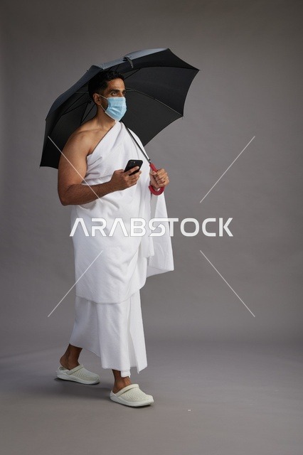 A Saudi Arabian Gulf pilgrim in Ihram clothing holding an umbrella and wearing a mask, using a mobile phone, performing the rituals of Hajj and Umrah, full body, gray background.