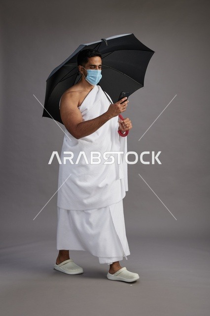 حاج  عربي سعودي خليجي بملابس الإحرام يحمل مظلة ويرتدي كمامة, استخدام الهاتف المحمول , اداء مناسك الحج والعمره, كامل الجسم, خلفيه رماديه
