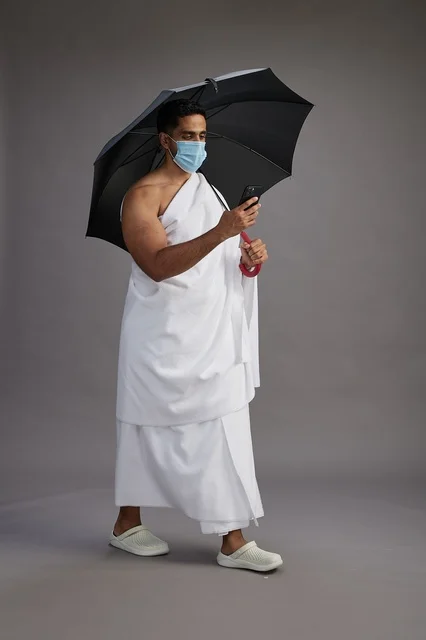A Saudi Arabian Gulf pilgrim in Ihram clothing holding an umbrella and wearing a mask, using a mobile phone, performing the rituals of Hajj and Umrah, full body, gray background.