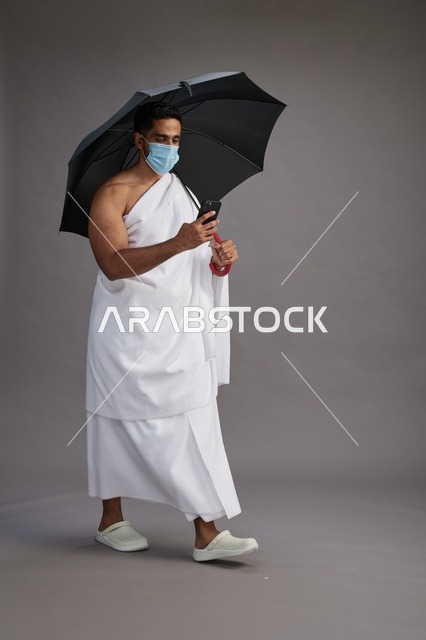 حاج  عربي سعودي خليجي بملابس الإحرام يحمل مظلة ويرتدي كمامة, استخدام الهاتف المحمول , اداء مناسك الحج والعمره, كامل الجسم, خلفيه رماديه