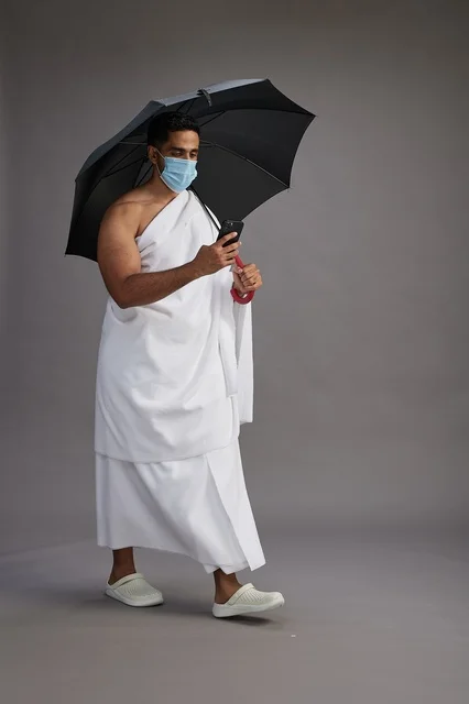 حاج  عربي سعودي خليجي بملابس الإحرام يحمل مظلة ويرتدي كمامة, استخدام الهاتف المحمول , اداء مناسك الحج والعمره, كامل الجسم, خلفيه رماديه