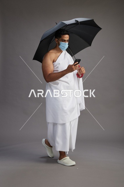 حاج  عربي سعودي خليجي بملابس الإحرام يحمل مظلة ويرتدي كمامة, استخدام الهاتف المحمول , اداء مناسك الحج والعمره, كامل الجسم, خلفيه رماديه