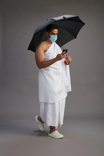 حاج  عربي سعودي خليجي بملابس الإحرام يحمل مظلة ويرتدي كمامة, استخدام الهاتف المحمول , اداء مناسك الحج والعمره, كامل الجسم, خلفيه رماديه