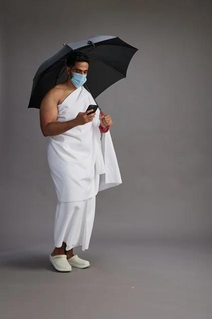 A Saudi Arabian Gulf pilgrim in Ihram clothing holding an umbrella and wearing a mask, using a mobile phone, performing the rituals of Hajj and Umrah, full body, gray background.