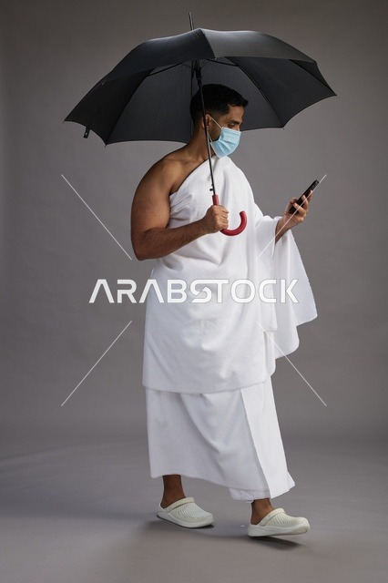 حاج  عربي سعودي خليجي بملابس الإحرام يحمل مظلة ويرتدي كمامة, استخدام الهاتف المحمول , اداء مناسك الحج والعمره, كامل الجسم, خلفيه رماديه