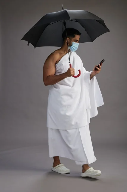 حاج  عربي سعودي خليجي بملابس الإحرام يحمل مظلة ويرتدي كمامة, استخدام الهاتف المحمول , اداء مناسك الحج والعمره, كامل الجسم, خلفيه رماديه
