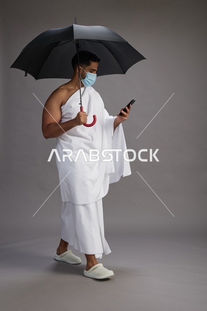 حاج  عربي سعودي خليجي بملابس الإحرام يحمل مظلة ويرتدي كمامة, استخدام الهاتف المحمول , اداء مناسك الحج والعمره, كامل الجسم, خلفيه رماديه