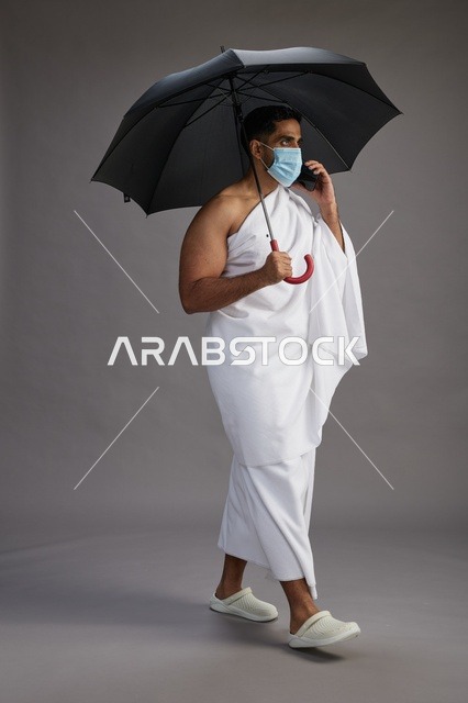 حاج  عربي سعودي خليجي بملابس الإحرام يحمل مظلة ويرتدي كمامة, استخدام الهاتف المحمول , اداء مناسك الحج والعمره, كامل الجسم, خلفيه رماديه