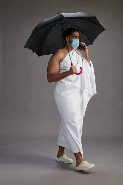 حاج عربي سعودي خليجي بملابس الإحرام يحمل مظلة ويرتدي كمامة, استخدام الهاتف المحمول , اداء مناسك الحج والعمره, كامل الجسم, خلفيه رماديه حاج عربي سعودي خليجي بملابس الإحرام يحمل مظلة ويرتدي كمامة, استخدام الهاتف المحمول , اداء مناسك الحج والعمره, كامل الجسم, خلفيه رماديه