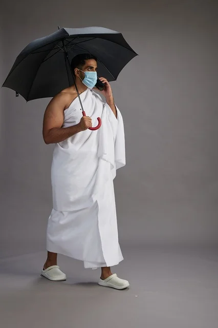 A Saudi Arabian Gulf pilgrim in Ihram clothing holding an umbrella and wearing a mask, using a mobile phone, performing the rituals of Hajj and Umrah, full body, gray background.