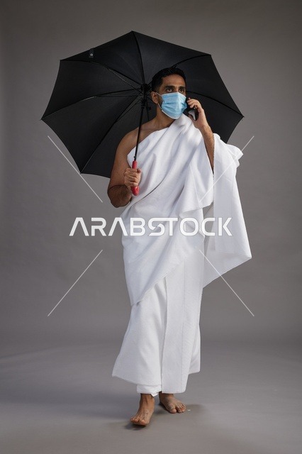 A Saudi Arabian Gulf pilgrim in Ihram clothing holding an umbrella and wearing a mask, using a mobile phone, performing the rituals of Hajj and Umrah, full body, gray background.
