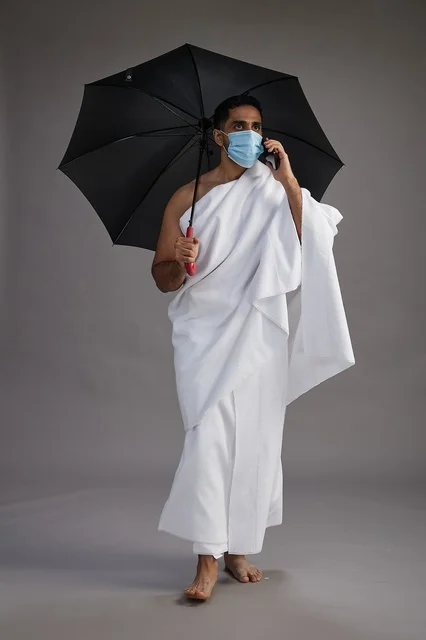 A Saudi Arabian Gulf pilgrim in Ihram clothing holding an umbrella and wearing a mask, using a mobile phone, performing the rituals of Hajj and Umrah, full body, gray background.