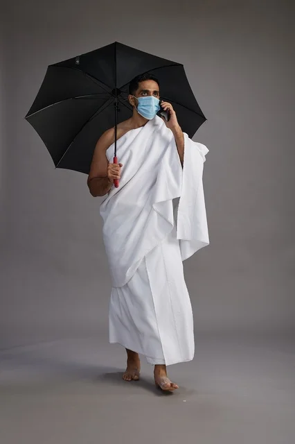 A Saudi Arabian Gulf pilgrim in Ihram clothing holding an umbrella and wearing a mask, using a mobile phone, performing the rituals of Hajj and Umrah, full body, gray background.