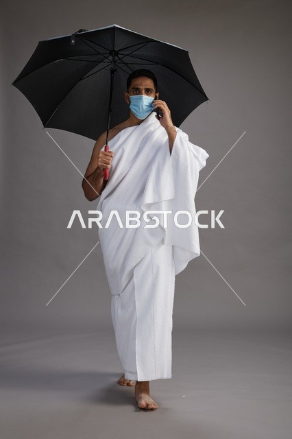 حاج  عربي سعودي خليجي بملابس الإحرام يحمل مظلة ويرتدي كمامة, استخدام الهاتف المحمول , اداء مناسك الحج والعمره, كامل الجسم, خلفيه رماديه