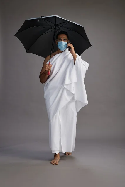 A Saudi Arabian Gulf pilgrim in Ihram clothing holding an umbrella and wearing a mask, using a mobile phone, performing the rituals of Hajj and Umrah, full body, gray background.