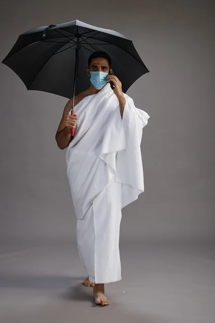 A Saudi Arabian Gulf pilgrim in Ihram clothing holding an umbrella and wearing a mask, using a mobile phone, performing the rituals of Hajj and Umrah, full body, gray background.