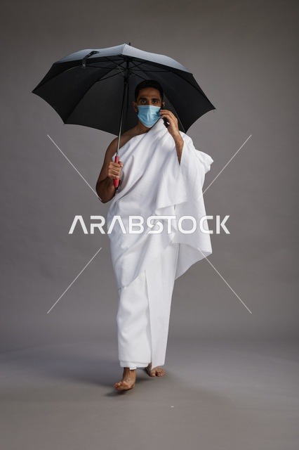 حاج  عربي سعودي خليجي بملابس الإحرام يحمل مظلة ويرتدي كمامة, استخدام الهاتف المحمول , اداء مناسك الحج والعمره, كامل الجسم, خلفيه رماديه