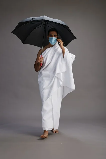 حاج  عربي سعودي خليجي بملابس الإحرام يحمل مظلة ويرتدي كمامة, استخدام الهاتف المحمول , اداء مناسك الحج والعمره, كامل الجسم, خلفيه رماديه