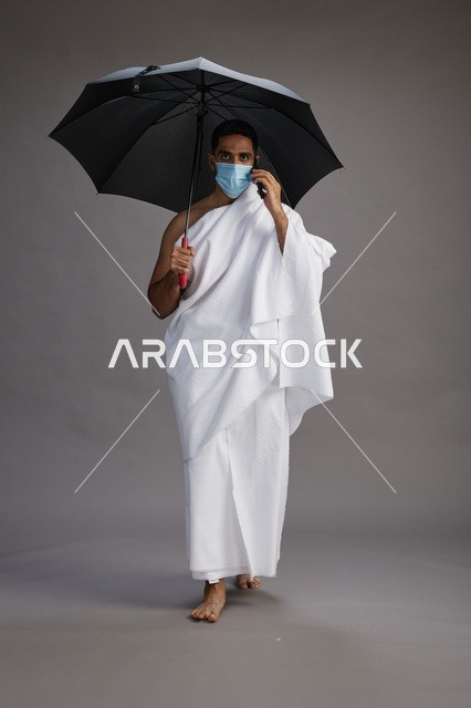 حاج  عربي سعودي خليجي بملابس الإحرام يحمل مظلة ويرتدي كمامة, استخدام الهاتف المحمول , اداء مناسك الحج والعمره, كامل الجسم, خلفيه رماديه