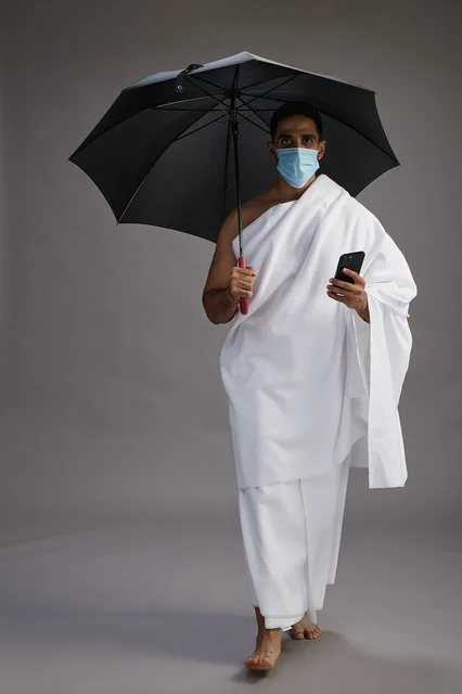 حاج  عربي سعودي خليجي بملابس الإحرام يحمل مظلة ويرتدي كمامة, استخدام الهاتف المحمول , اداء مناسك الحج والعمره, كامل الجسم, خلفيه رماديه
