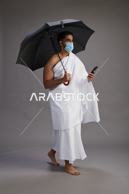 حاج  عربي سعودي خليجي بملابس الإحرام يحمل مظلة ويرتدي كمامة, استخدام الهاتف المحمول , اداء مناسك الحج والعمره, كامل الجسم, خلفيه رماديه