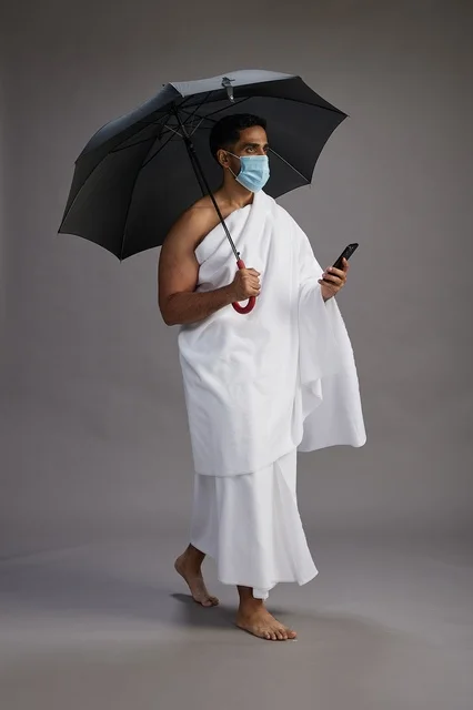 A Saudi Arabian Gulf pilgrim in Ihram clothing holding an umbrella and wearing a mask, using a mobile phone, performing the rituals of Hajj and Umrah, full body, gray background.