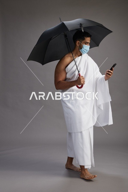 A Saudi Arabian Gulf pilgrim in Ihram clothing holding an umbrella and wearing a mask, using a mobile phone, performing the rituals of Hajj and Umrah, full body, gray background.