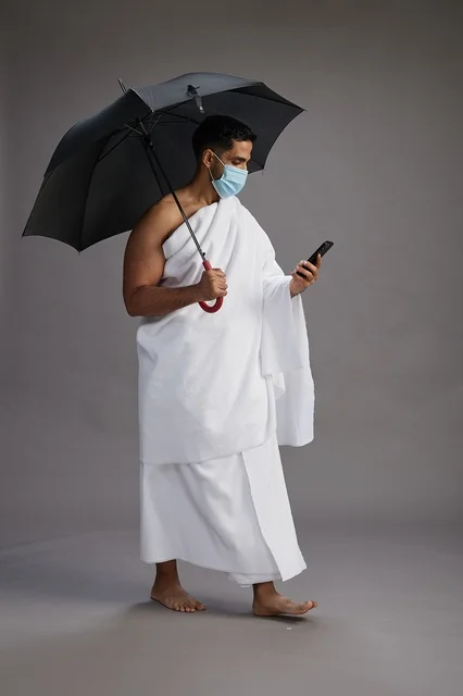 A Saudi Arabian Gulf pilgrim in Ihram clothing holding an umbrella and wearing a mask, using a mobile phone, performing the rituals of Hajj and Umrah, full body, gray background.