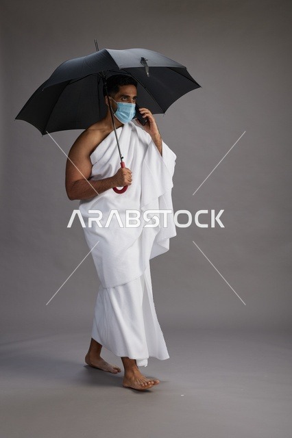 حاج  عربي سعودي خليجي بملابس الإحرام يحمل مظلة ويرتدي كمامة, استخدام الهاتف المحمول , اداء مناسك الحج والعمره, كامل الجسم, خلفيه رماديه