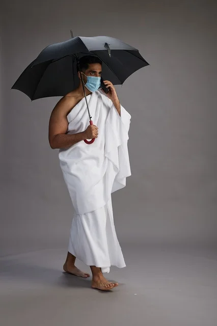 حاج  عربي سعودي خليجي بملابس الإحرام يحمل مظلة ويرتدي كمامة, استخدام الهاتف المحمول , اداء مناسك الحج والعمره, كامل الجسم, خلفيه رماديه