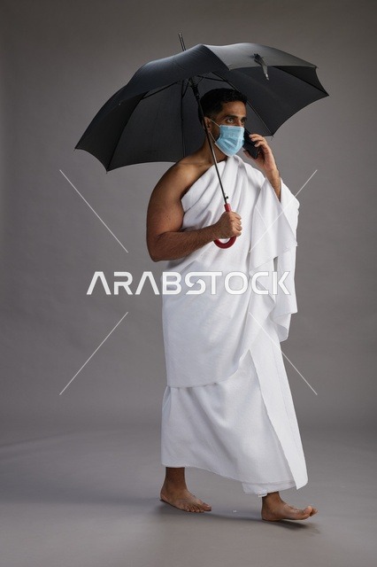 حاج  عربي سعودي خليجي بملابس الإحرام يحمل مظلة ويرتدي كمامة, استخدام الهاتف المحمول , اداء مناسك الحج والعمره, كامل الجسم, خلفيه رماديه