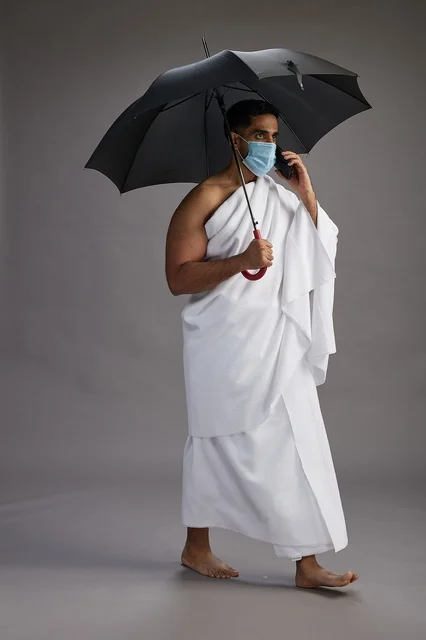حاج  عربي سعودي خليجي بملابس الإحرام يحمل مظلة ويرتدي كمامة, استخدام الهاتف المحمول , اداء مناسك الحج والعمره, كامل الجسم, خلفيه رماديه