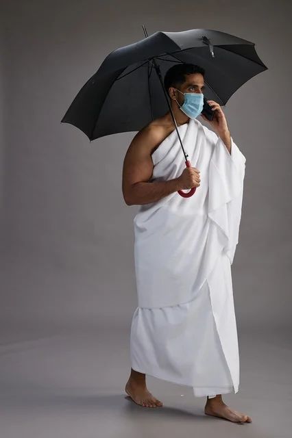 A Saudi Arabian Gulf pilgrim in Ihram clothing holding an umbrella and wearing a mask, using a mobile phone, performing the rituals of Hajj and Umrah, full body, gray background.