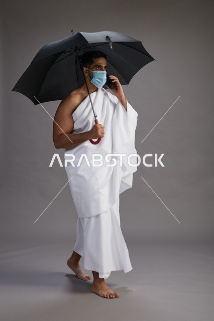 حاج  عربي سعودي خليجي بملابس الإحرام يحمل مظلة ويرتدي كمامة, استخدام الهاتف المحمول , اداء مناسك الحج والعمره, كامل الجسم, خلفيه رماديه