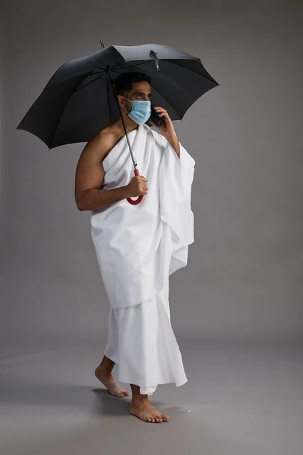 A Saudi Arabian Gulf pilgrim in Ihram clothing holding an umbrella and wearing a mask, using a mobile phone, performing the rituals of Hajj and Umrah, full body, gray background.