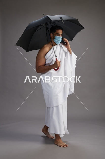 حاج  عربي سعودي خليجي بملابس الإحرام يحمل مظلة ويرتدي كمامة, استخدام الهاتف المحمول , اداء مناسك الحج والعمره, كامل الجسم, خلفيه رماديه