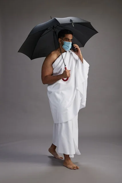 حاج  عربي سعودي خليجي بملابس الإحرام يحمل مظلة ويرتدي كمامة, استخدام الهاتف المحمول , اداء مناسك الحج والعمره, كامل الجسم, خلفيه رماديه