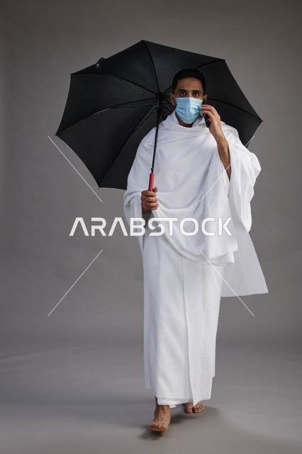 حاج  عربي سعودي خليجي بملابس الإحرام يحمل مظلة ويرتدي كمامة, استخدام الهاتف المحمول , اداء مناسك الحج والعمره, كامل الجسم, خلفيه رماديه
