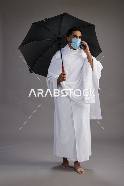 حاج  عربي سعودي خليجي بملابس الإحرام يحمل مظلة ويرتدي كمامة, استخدام الهاتف المحمول , اداء مناسك الحج والعمره, كامل الجسم, خلفيه رماديه