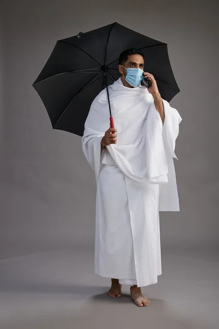 حاج  عربي سعودي خليجي بملابس الإحرام يحمل مظلة ويرتدي كمامة, استخدام الهاتف المحمول , اداء مناسك الحج والعمره, كامل الجسم, خلفيه رماديه